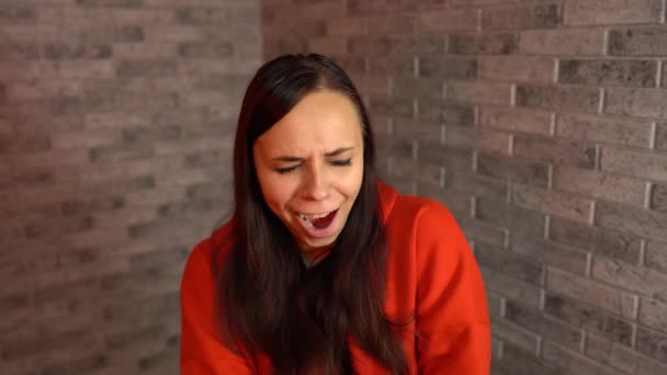 Een mooie jonge vrouw in een rode hoodie geeuwt van vermoeidheid op een bakstenen achtergrond. — Stockvideo