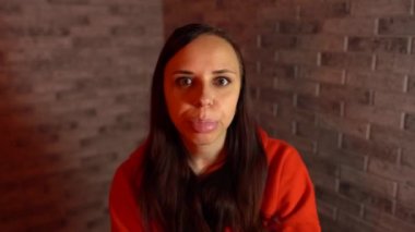 Kırmızı kapüşonlu genç bir kadın, bir tuğla arka planda kameraya bakarak dilini aşağılıyor..