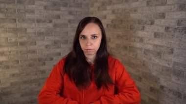 Kırmızı kapüşonlu genç bir kadın, bir tuğla arka planda kameraya bakarak dilini aşağılıyor..