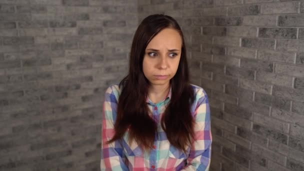 Een mooie jonge vrouw in een geruit shirt is in een slechte bui. Een jonge vrouw toont een verdriet op een bakstenen achtergrond. — Stockvideo