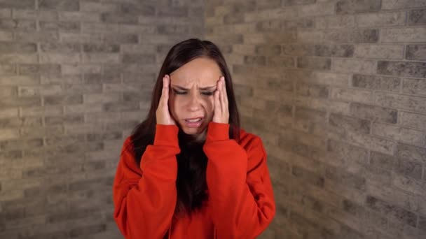 Uma mulher nova em um hoodie vermelho está prendendo suas mãos em temples na dor em um fundo do tijolo. — Vídeo de Stock
