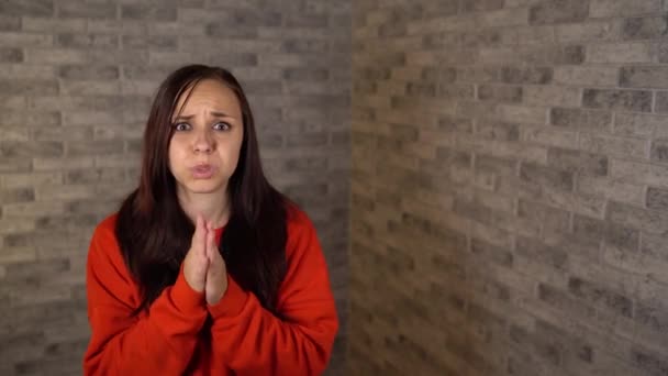 Примхлива молода жінка в червоному светрі благає вираження. Брюнетка пестить губи і тримає руки в молитві . — стокове відео