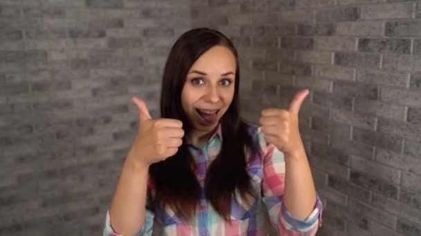 A beautiful emotional woman in a plaid shirt laughs happily and shows thumbs up. A woman shows a gesture of approval on a brick background. The concept of like or dislike. — Stock Video