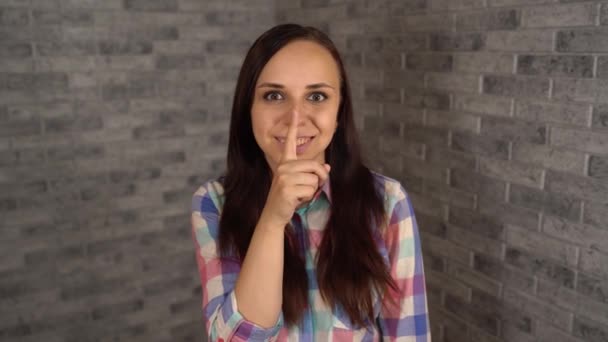 Een mooie emotionele vrouw in een geruite overhemd doet het stilzwijgen gebaar met wijsvinger naar haar lippen op een grijze bakstenen achtergrond. Het begrip stilte. — Stockvideo