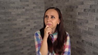 Ekose gömlekli genç bir kadın gri tuğlalı arka plandaki bir şeyi düşünüyor. Düşünceli bir esmer etrafına bakıyor ve elini çenesinde tutuyor..