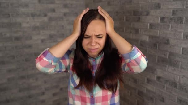 A young woman in a plaid shirt grabs her head with hands in pain on a brick background. A woman has a headache. — Stock Video