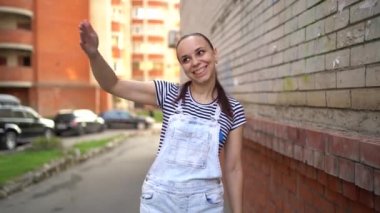 Gündelik giysiler içinde genç bir kadın geniş bir şekilde gülümsüyor ve elini sallayarak birine sesleniyor..