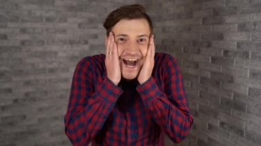 Ekose gömlekli genç yakışıklı bir adam ağzını açıyor ve elleri yanaklarında gri tuğla bir arka planda hoş bir sürpriz yapıyor..