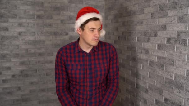 A young handsome man in Santa Clause hat is being in bad mood. A young man is showing a sadness on a gray brick background. — Stock Video