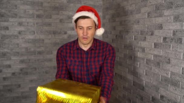 Un jeune bel homme portant le chapeau Santa Clause reçoit les cadeaux enveloppés. Un homme heureux tient des cadeaux enveloppés et regarde la caméra sur un mur de briques grises. — Video