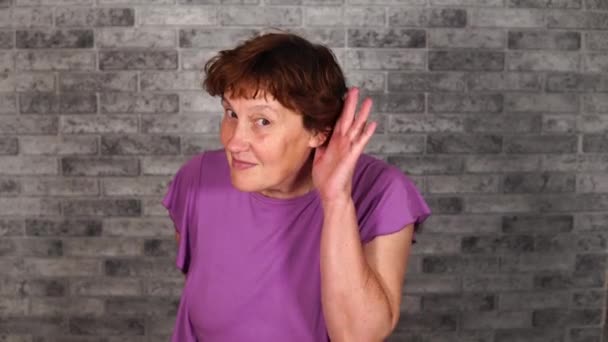 A beautiful grown woman in casual clothes is holding hand at her ear and trying to overhear conversation on a gray brick background. A curious mature woman is smiling broadly and spying on someone — Stock Video