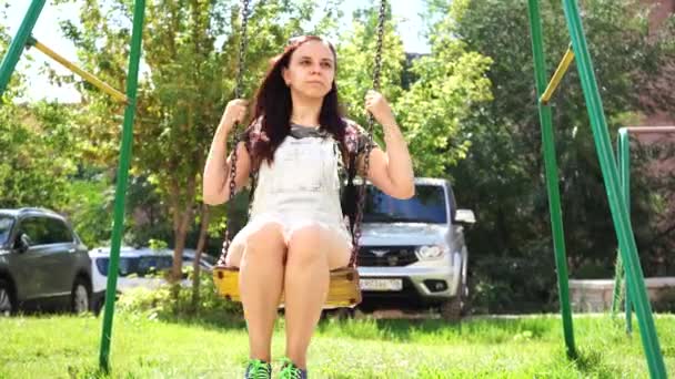 Eine junge Frau in legerer Kleidung schwingt auf einer Schaukel auf dem Spielplatz. — Stockvideo