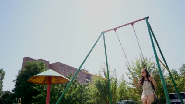 Una joven con ropa casual se balancea en un columpio en el patio de recreo . — Vídeo de stock