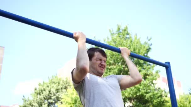 A young handsome man in casual clothes jokingly is doing pull up exercise on horizontal bar. The concept of humor, jokes and sports. — Stock Video