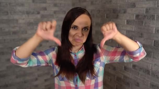 A beautiful emotional woman in a plaid shirt shows thumbs down. A woman shows a gesture of disapproval on a brick background. The concept of like or dislike. – Stock-video