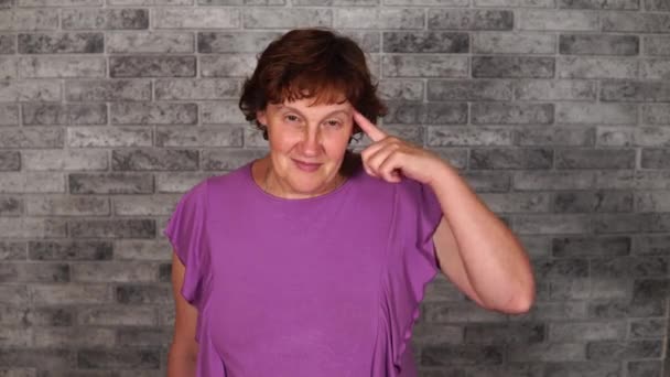 A beautiful mature woman in casual clothes is twisting at her temple on a gray brick background. — Stock Video