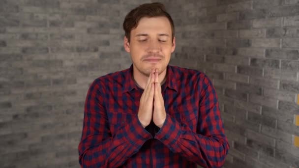 Молодий красивий чоловік у сорочці тримає руки в молитві на сірому цегляному фоні . — стокове відео