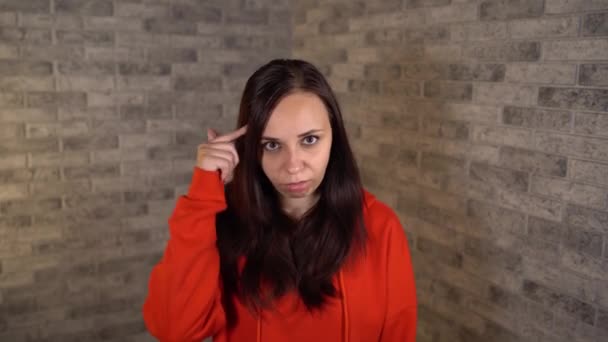 Eine schöne junge Frau in rotem Kapuzenpulli dreht sich vor grauem Backsteinhintergrund um ihre Schläfe. — Stockvideo