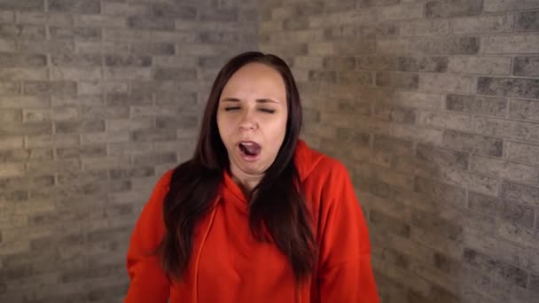 A beautiful young woman in a red hoodie yawns from fatigue on a brick background. – Stock-video