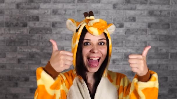 Uma mulher engraçada em um grande kigurumi de girafa está sorrindo e mostrando polegares para cima. Uma mulher mostra um gesto de aprovação em um fundo de tijolo cinza. O conceito de Halloween, festa de pijama e animador para — Vídeo de Stock