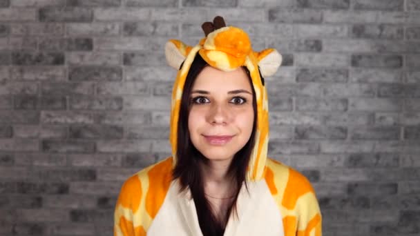 Une femme drôle dans un grand kigurumi de girafe est agréablement surprise avec les mains sur ses joues sur un fond de brique grise. Le concept d'Halloween, de fête de pajama et d'animateur pour les fêtes d'enfants. — Video