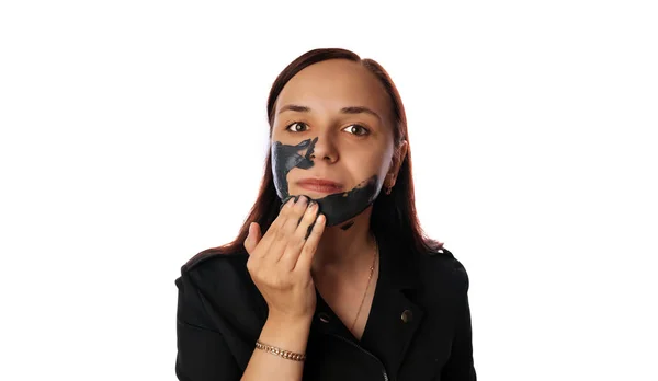 Eine junge Frau setzt dem Gesicht auf weißem Hintergrund eine schwarze Maske auf. Das Konzept von gesundem Lebensstil, Schönheit und Körperpflege. — Stockfoto