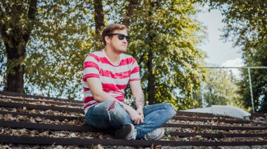 Gündelik giysiler içinde genç yakışıklı bir adam parktaki merdivenlerde oturuyor ve etrafa bakıyor..