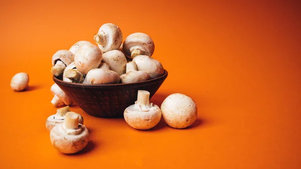 Mushrooms in a wooden bowl on an orange background. The small white champignon in a plate and scattered near it. — Stock Image Mushrooms in a wooden bowl on an orange background. The small white champignon in a plate and scattered near it. Stock Picture