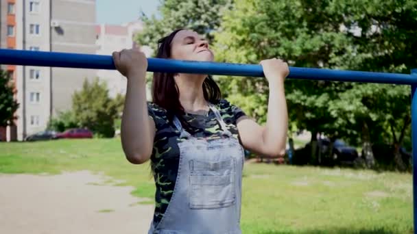 Eine junge Frau in lässiger Kleidung macht scherzhaft eine Klimmzugübung am Reck. Das Konzept von Humor, Witzen und Sport. — Stockvideo