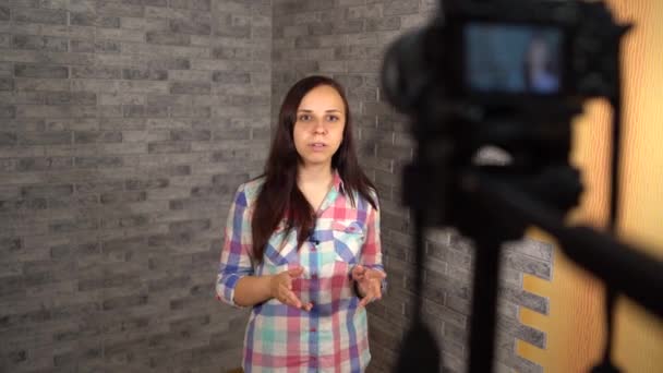 Uma jovem mulher em uma camisa de xadrez está falando na câmera em um fundo de tijolo. Uma mulher feliz está falando na frente da câmera para vlog. — Vídeo de Stock
