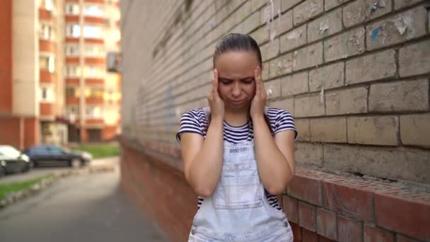 Een jonge vrouw in casual kleding houdt haar handen op tempels in pijn in de straat. — Stockvideo