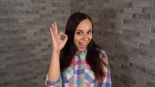Een mooie jonge vrouw in een geruit shirt glimlacht en vertoont tekenen van OK op een grijze bakstenen achtergrond. — Stockvideo