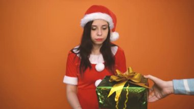 Noel Baba kostümüyle gülümseyen genç bir kadın turuncu arka planda hediyeler tutuyor. Mutlu Noeller ve mutlu yıllar..