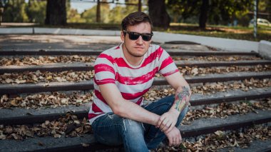 Gündelik giysiler içinde genç yakışıklı bir adam parktaki merdivenlerde oturuyor ve etrafa bakıyor..