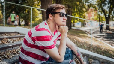 Gündelik giysiler içinde genç yakışıklı bir adam parktaki merdivenlerde oturuyor ve etrafa bakıyor..