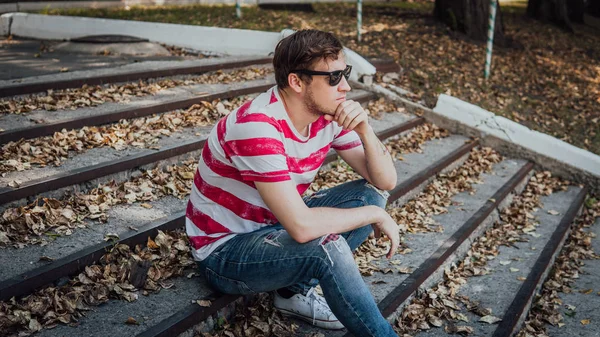 Um jovem bonito homem em roupas casuais está sentado nos degraus do parque e olhando ao redor. — Fotografia de Stock