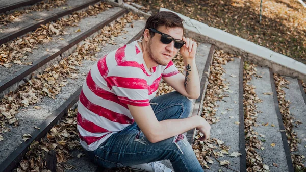 Gündelik giysiler içinde genç yakışıklı bir adam parktaki merdivenlerde oturuyor ve etrafa bakıyor..