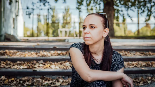 Une femme pensive vêtue d'un maillot de ville noir est assise sur les marches avec des feuilles tombées dans le parc. — Photo