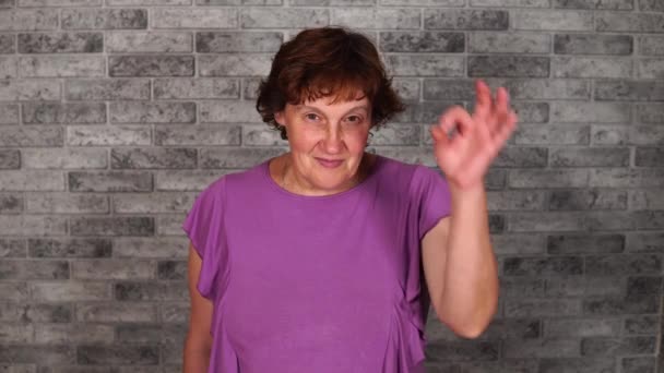 A beautiful mature woman in casual clothes is smiling and showing sign of OK on a gray brick background. — Stock Video