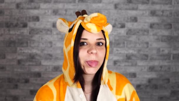 Una donna divertente in un grande kigurumi di giraffa sta sorridendo e mostrando la sua lingua guardando la fotocamera su uno sfondo di mattoni grigi. — Video Stock