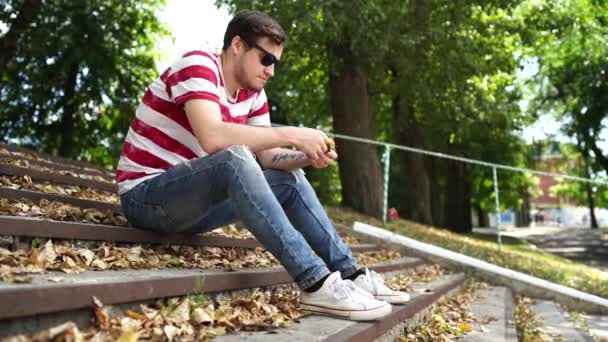 Um jovem bonito homem em roupas casuais está sentado nos degraus do parque e olhando ao redor. — Vídeo de Stock