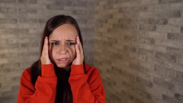 Egy fiatal nő piros kapucnis pulcsiban a kezét a halántékán tartja fájdalommal a tégla hátterében.. — Stock videók