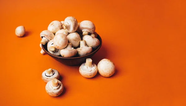 Mushrooms in a wooden bowl on an orange background. The small white champignon in a plate and scattered near it. — Stock Image Mushrooms in a wooden bowl on an orange background. The small white champignon in a plate and scattered near it. Stock Photo