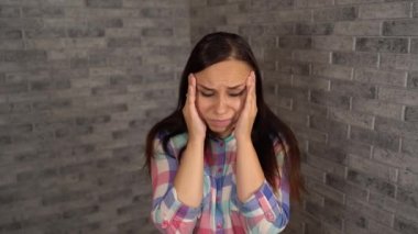 Ekose gömlekli genç ve güzel bir adam gri tuğlalı arka planda acı içinde ellerini şakaklarına koyuyor..