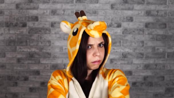 Vtipná žena ve velké žirafí kigurumi má špatnou náladu. Krásná emocionální žena v pyžamu ukazuje urážku na cihlovém pozadí. — Stock video