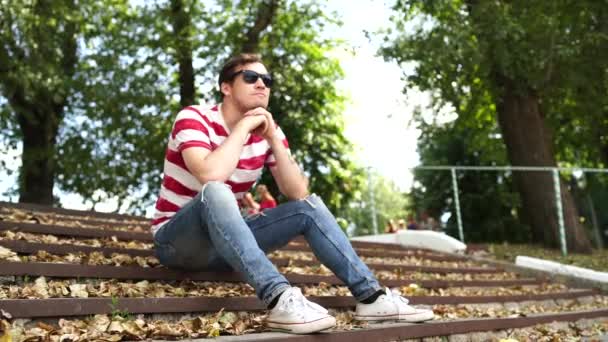 Un joven guapo con ropa casual está sentado en los escalones del parque y mirando a su alrededor.. — Vídeos de Stock