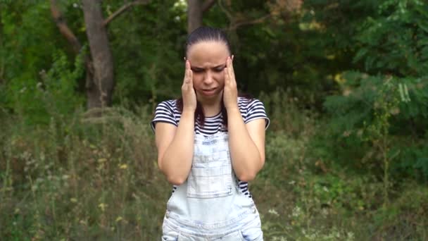 Una joven vestida con ropa ocasional está tendiendo las manos sobre los templos que sufren en el bosque.. — Vídeos de Stock