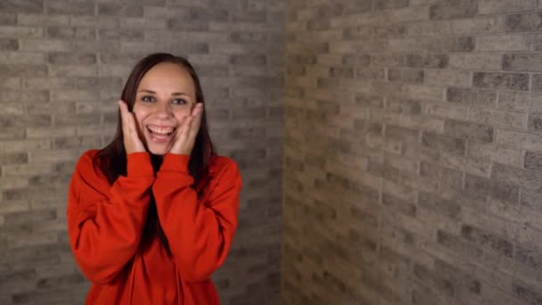 Een mooie emotionele vrouw in een rode hoodie is blij verrast met handen op haar wangen. — Stockvideo