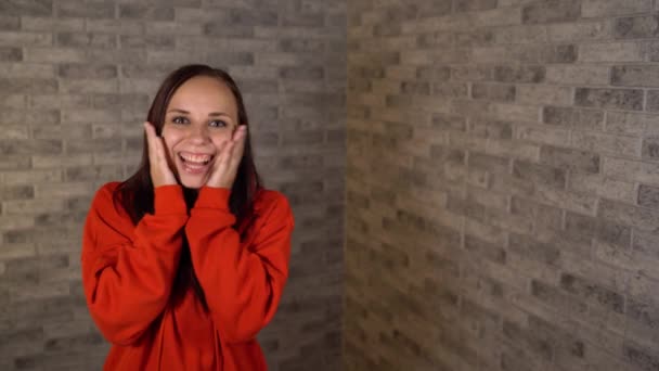 Une belle femme affective dans un hoodie rouge est agréablement surprise avec les mains sur ses joues. — Video