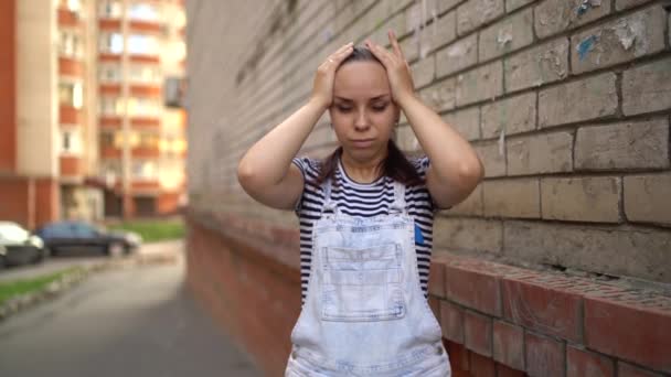 Młoda kobieta w luźnych ciuchach trzyma ręce na skroniach w bólu na ulicy.. — Wideo stockowe